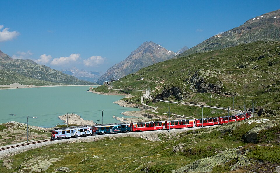 Two RhB ABe 4/4III motor coaches in multiple unit with a Bernina Express train on the Bernina line, passing Lago Bianco with Ospizio Bernina railway station in the background.